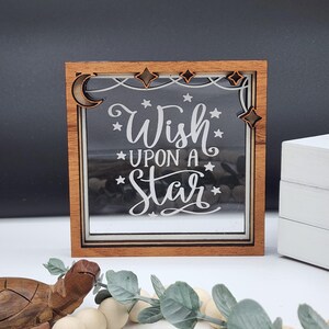 Peut inclure: Un cadre en bois avec un insert en verre présentant le texte "Wish upon a star" en lettres blanches. Le cadre est décoré d'un croissant de lune et d'étoiles.