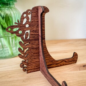 May include: A wooden display stand with a butterfly design. The stand is made of dark brown wood and has a natural wood grain finish. The stand is designed to hold a plate or other decorative object.