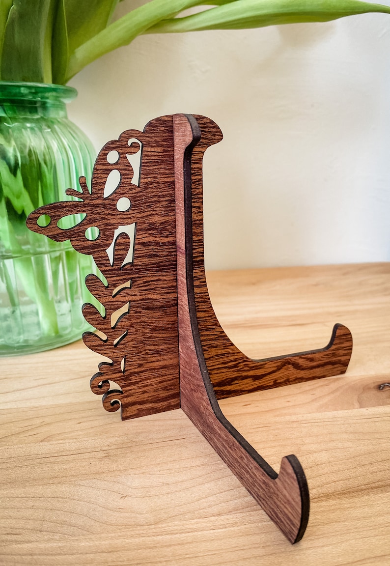 May include: A wooden display stand with a butterfly design. The stand is made of dark brown wood and has a natural wood grain finish. The stand is designed to hold a plate or other decorative object.