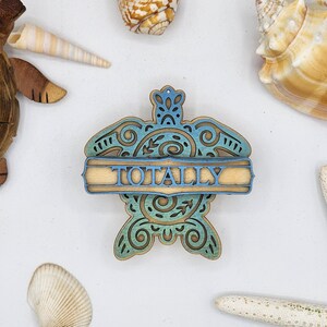 May include: A wooden turtle ornament with a blue and green gradient paint finish. The turtle has a decorative design and a banner that reads "TOTALLY".