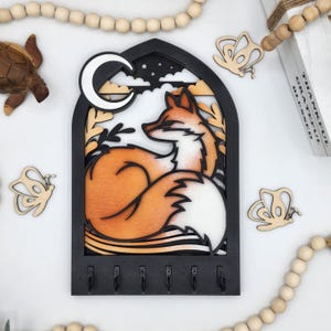 May include: A black wooden key holder shaped like a window with a fox curled up inside. The fox is orange and white with a crescent moon above it. There are five hooks on the bottom of the key holder.