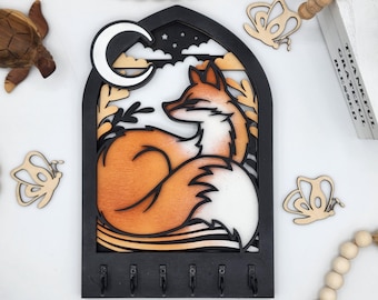 May include: A black wooden key holder shaped like a window with a fox curled up inside. The fox is orange and white with a crescent moon above it. There are five hooks on the bottom of the key holder.
