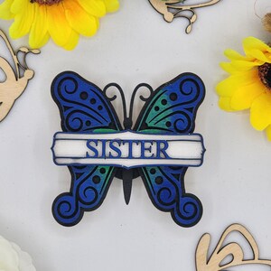 May include: Wooden butterfly cutout with a blue and green gradient paint finish. The butterfly has a white banner across the middle with the word "SISTER" written in black.