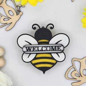 May include: Wooden bee cutout with black and yellow stripes and white wings. The bee is holding a sign that says "WELCOME" in black letters.