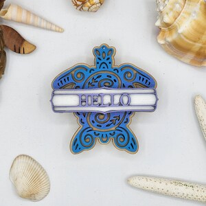 May include: A blue and gold wooden turtle ornament with a white banner that says "HELLO".
