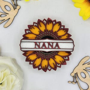 May include: A wooden sunflower with a red and white banner that says "NANA". The sunflower is yellow and has a red outline. The banner is white with red lettering.