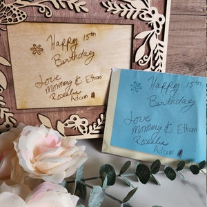May include: A wooden picture frame with a floral design and a handwritten message that says "Happy 15th Birthday Love Mommy & Ethan Rosalie Adam". There is a blue sticky note with the same message on it.