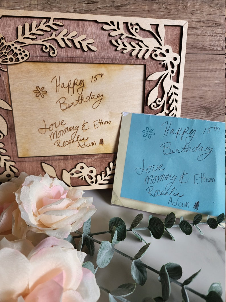 May include: A wooden picture frame with a floral design and a handwritten message that says "Happy 15th Birthday Love Mommy & Ethan Rosalie Adam". There is a blue sticky note with the same message on it.
