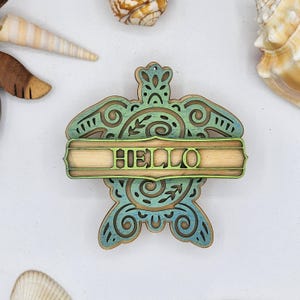 May include: A wooden turtle ornament with a green and blue gradient paint finish. The turtle has intricate details and a banner that reads "HELLO".