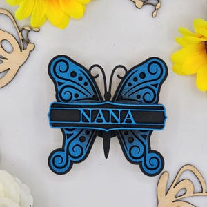 May include: A blue wooden butterfly with a black banner that says "NANA" on it. The butterfly has intricate details and a gradient of blue.