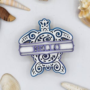 May include: A white wooden turtle ornament with blue accents and a white banner that says "HELLO" in purple lettering.