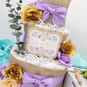 May include: A three-tier diaper cake decorated with burlap, purple ribbon, and gold and peach flowers. The top tier has a sign that says "Baby in Bloom".