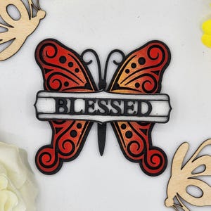 May include: Wooden butterfly cutout with a red and orange gradient, black details, and the word "BLESSED" written on a white banner.
