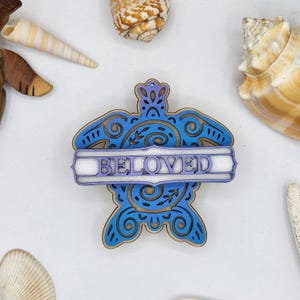 May include: A wooden turtle-shaped ornament with the word "BELOVED" in a white frame. The ornament features blue and purple layered details with decorative swirls and floral accents. Seashells are scattered around the ornament.