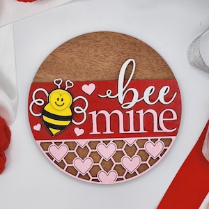 May include: A round wooden sign with a red and pink honeycomb pattern. The sign says "bee mine" in white lettering and features a cartoon bee with a yellow face and black and yellow stripes.