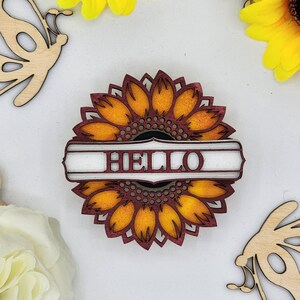 May include: A wooden sunflower with a red and white sign that says "HELLO" in the centre. The sunflower is yellow and orange with a red outline.