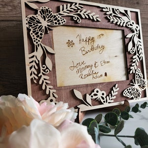 May include: A wooden picture frame with a cutout design of butterflies and leaves. The frame has a light brown stain and a dark brown wood insert. The frame has a message inside that reads "Happy 15th Birthday Love Mommy & Ethan Rosalie Adam"