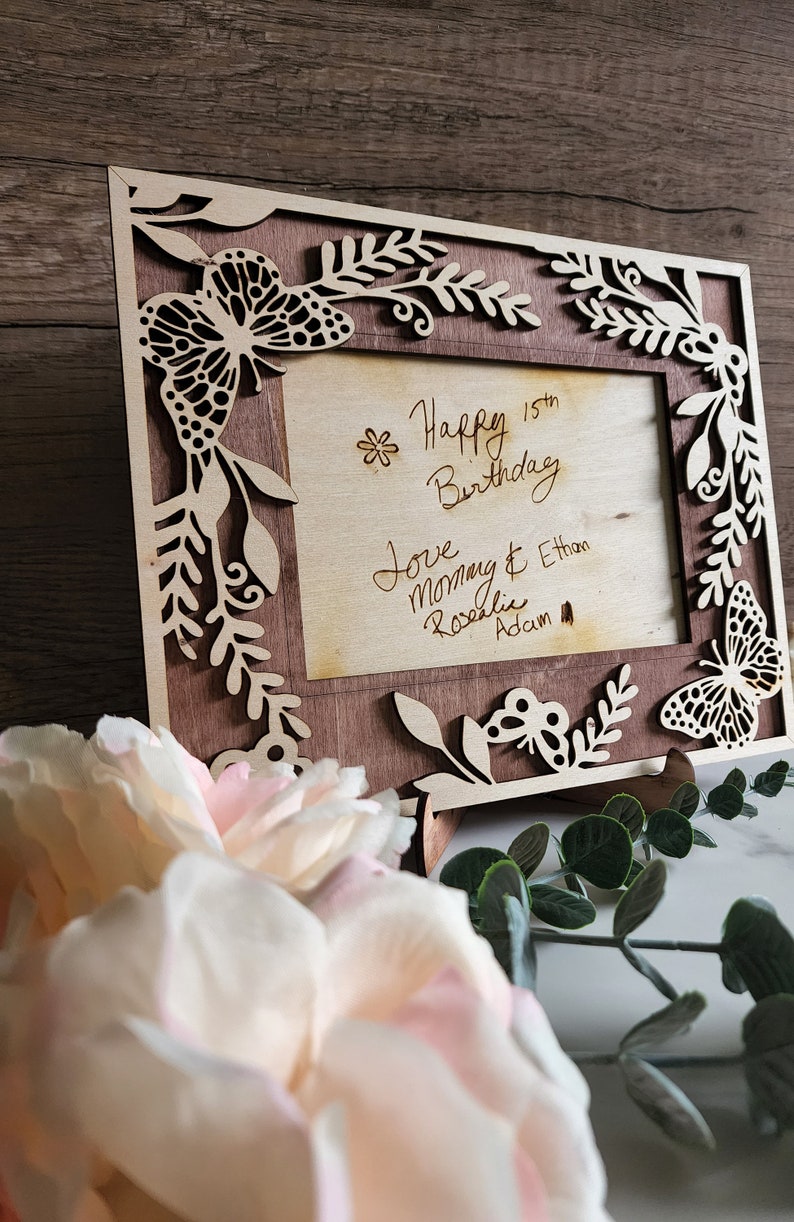 May include: A wooden picture frame with a cutout design of butterflies and leaves. The frame has a light brown stain and a dark brown wood insert. The frame has a message inside that reads "Happy 15th Birthday Love Mommy & Ethan Rosalie Adam"