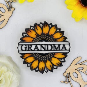 May include: A wooden cutout of a sunflower with a white banner that reads "GRANDMA". The sunflower is yellow and black with a brown centre.