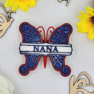 May include: A wooden butterfly with red and blue paint and a white banner that says "NANA".