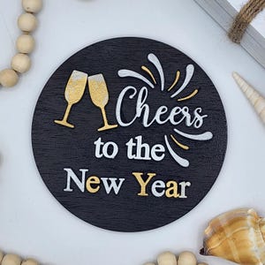 May include: Circular black wooden sign with the text "Cheers to the New Year" in white and gold. Two gold champagne glasses are featured. A decorative item for New Year celebrations.