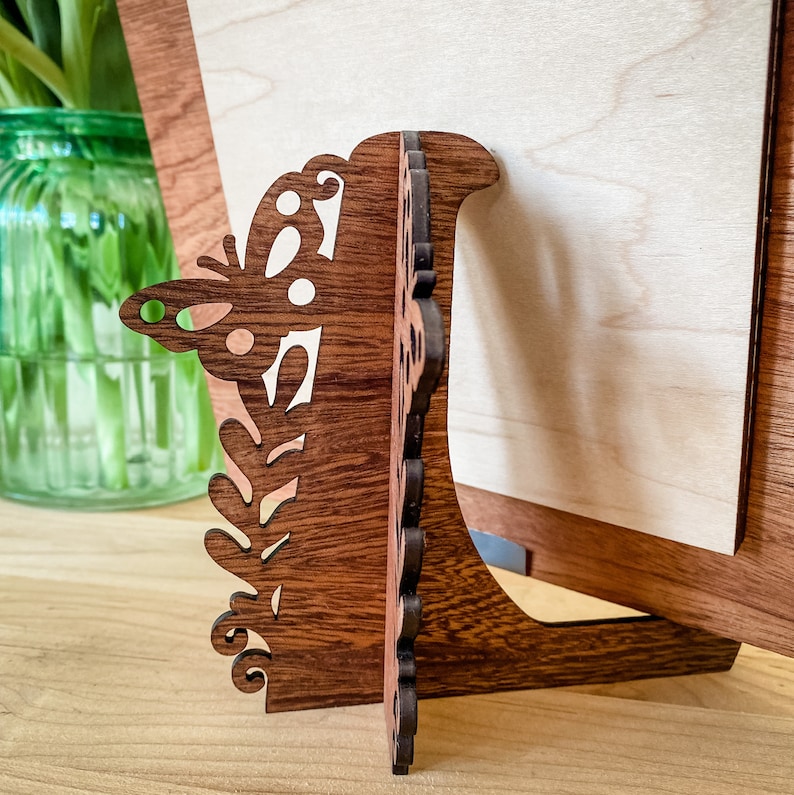 May include: A wooden picture frame with a decorative butterfly design. The frame is made of dark wood and has a light wood backing. The frame is standing on a table.