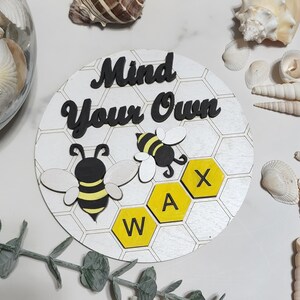 Peut inclure: Une pancarte en bois blanc avec un motif en nid d'abeille et le texte "Mind Your Own Wax" en lettres noires. Deux abeilles noires et jaunes sont sur la pancarte, et les lettres "W", "A" et "X" sont sur le nid d'abeille.