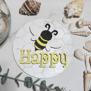 Peut inclure: Une pancarte en bois blanc avec un motif en nid d'abeille et le mot "Happy" en lettres jaunes. Une abeille noire et jaune avec des ailes est centrée sur la pancarte.