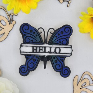 May include: A blue and black wooden butterfly with a white banner that says "HELLO".