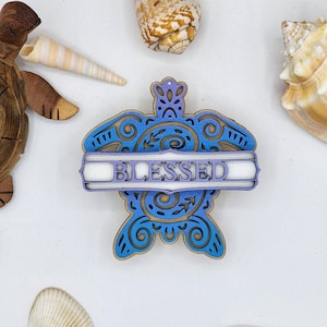 May include: A blue and purple wooden turtle-shaped ornament with the word "BLESSED" in white. The ornament has intricate cut-out designs and is surrounded by seashells and a wooden turtle figurine.