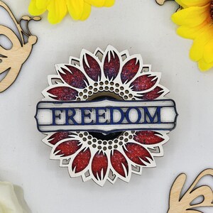 May include: Wooden cutout of a red, white and blue sunflower with the word "FREEDOM" in a blue banner across the centre.