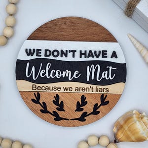 May include: Round wooden sign with the text "WE DON'T HAVE A Welcome Mat Because we aren't liars". The sign has a brown, white, black, and tan color scheme with a decorative leaf design.
