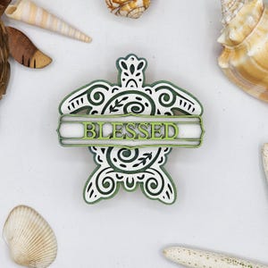 May include: A wooden turtle ornament with a layered green and white design. The word "BLESSED" is displayed on the shell. The ornament is surrounded by seashells on a white background.