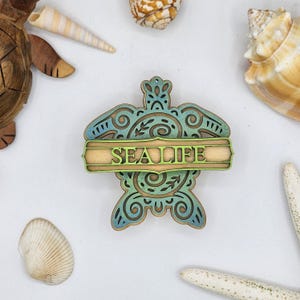 May include: A wooden cutout of a sea turtle with a green and blue gradient. The turtle is decorated with intricate swirls and the word "SEALIFE" is written on a banner across the turtle's shell.