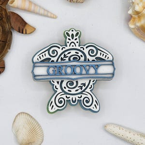 May include: White wooden turtle cutout with a blue banner that says "GROOVY" in white letters. The turtle has a detailed design with black swirls and lines.
