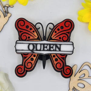 May include: A wooden butterfly with red and orange wings and black details. The butterfly has a white banner across its wings with the word "QUEEN" in black lettering.