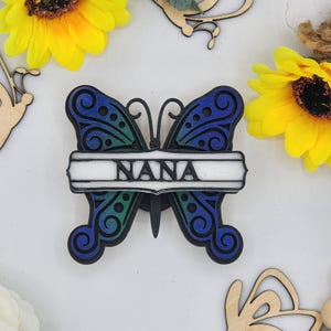 May include: A wooden butterfly decoration with the word "NANA" in white. The butterfly has blue and green wings with decorative patterns. Yellow sunflowers and other wooden cutouts are in the background.