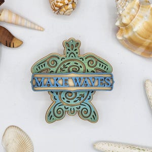May include: Wooden turtle-shaped ornament with the words "MAKE WAVES" in blue. The ornament has a layered design with teal and blue accents. Seashells surround the ornament on a white background. The ornament is likely for coastal or beach-themed decor.