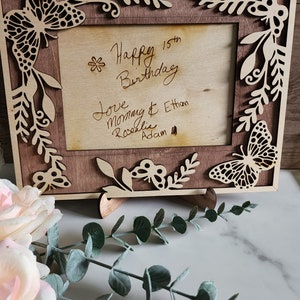 May include: A wooden picture frame with a cutout butterfly design. The frame has a light brown stain and is decorated with a floral pattern. The frame is standing on a white surface. The text inside the frame reads "Happy 15th Birthday Love Mommy & Ethan Rosalie Adam"