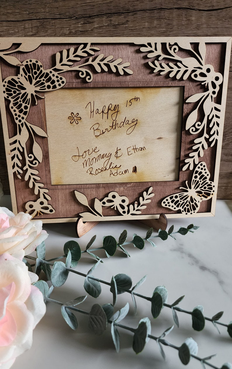 May include: A wooden picture frame with a cutout butterfly design. The frame has a light brown stain and is decorated with a floral pattern. The frame is standing on a white surface. The text inside the frame reads "Happy 15th Birthday Love Mommy & Ethan Rosalie Adam"