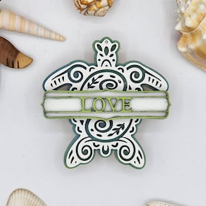 May include: A wooden turtle ornament with a green and blue swirl design. The turtle has a white banner across its shell with the word "LOVE" in green letters.