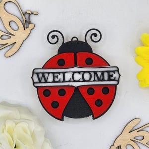 May include: Wooden ladybug welcome sign with red and black paint and white lettering. The sign says "WELCOME" and has a black background.
