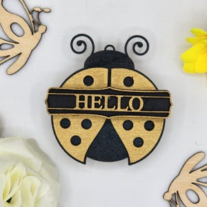 May include: Wooden ladybug ornament painted in black and gold. The ladybug has a black banner across its body with the word "HELLO" written in gold letters.