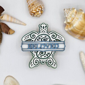 May include: A white wooden turtle ornament with a blue "BELOVED" banner. The turtle has a green and white swirling pattern.
