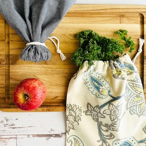 May include: A solid dark gray fabric produce bag tied with a white drawstring, a cream-colored fabric produce bag featuring a blue and green paisley floral pattern, and a single red apple, all arranged on a rectangular wooden cutting board.