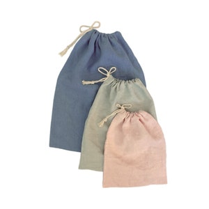 May include: Three drawstring pouches made of fabric in varying sizes and colors. The largest is slate blue, the medium is sage green, and the smallest is dusty pink. Each bag features a simple knotted cream-colored cord closure at the top.