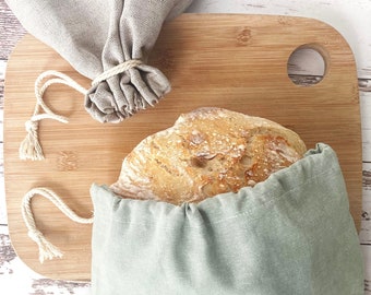 Peut inclure: Un sac à pain en tissu vert sauge avec une fermeture à cordon contenant une miche de pain croustillante, placé à côté d'un deuxième sac en tissu beige sur une planche à découper en bois. Les sacs sont en textile tissé avec des cordons.