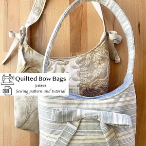 May include: Two quilted bow bags in neutral tones. One bag features a floral pattern, while the other has blue and white stripes. Both bags have bow accents and matching straps. The text on the image reads "Quilted Bow Bags, 3 sizes, Sewing pattern and tutorial."