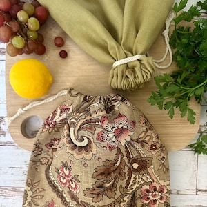 May include: Two drawstring produce bags, one with a floral paisley pattern and the other solid olive green, sit on a wooden cutting board. A lemon, grapes, and parsley are also on the board. The bags are designed for reusable grocery shopping.