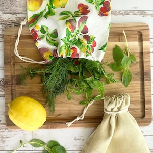 May include: A wooden cutting board with fresh herbs, a lemon, and two drawstring bags. One bag is filled with dill and mint, and the other is empty. The bags are made of linen and cotton. The bag with herbs has a white fabric with a colorful fruit pattern.